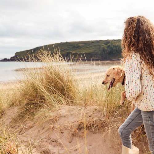 Girl Carrying Pet Dog Exploring Sand Dunes On Winter Beach Vacation